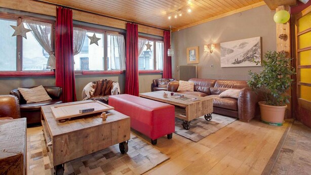 A cozy living room with a wooden ceiling, leather sofas, a red ottoman, and a large window with red curtains. - Hotel De La Poste (Bagnes)