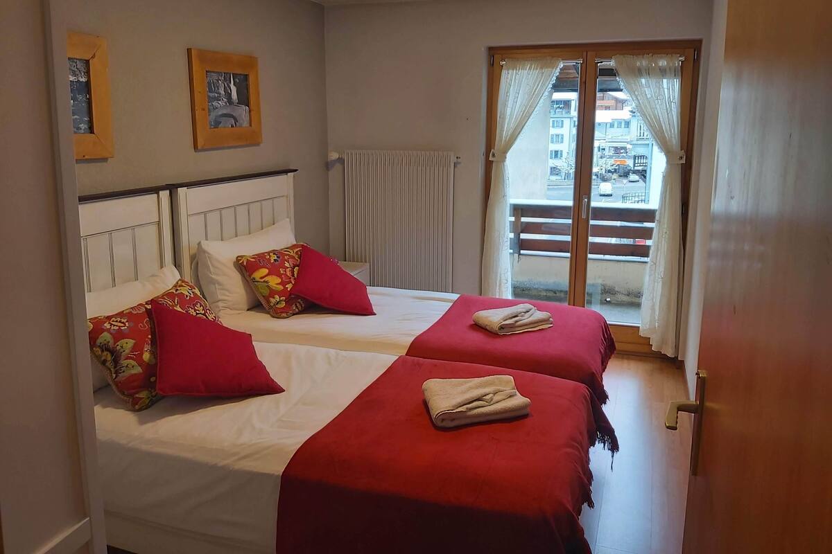 A hotel room with two beds, a window with curtains, a radiator, and a view of the outside.