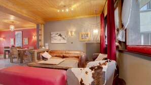 A cozy living room with a wooden ceiling, a red sofa, a leather couch, and a dining area with wooden furniture. - Hotel De La Poste (Bagnes)