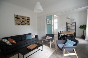 Un salon moderne avec un canapé gris foncé, une table basse en bois et deux fauteuils gris. Un luminaire suspendu blanc est accroché au-dessus du canapé, et un tableau est exposé au mur.