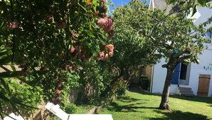 Um jardim com duas cadeiras de descanso brancas, uma árvore com flores cor-de-rosa e um edifício branco com uma janela azul.
