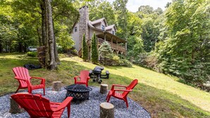 Una cabaña con chimenea de piedra, un balcón y una fogata rodeada de sillas Adirondack rojas.