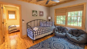 Un dormitorio acogedor con una cama, un sillón informal tipo bean bag color gris, un tapete estampado y una ventana con vistas a árboles.