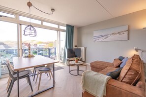 Een moderne woonkamer met een bruine leren bank, een eettafel met stoelen, een televisie en een groot raam met uitzicht op huizen en heuvels.