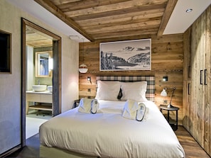 A bedroom with a wooden ceiling, a large bed with white linens, a mounted picture of a mountain landscape, and a nightstand with a lamp.