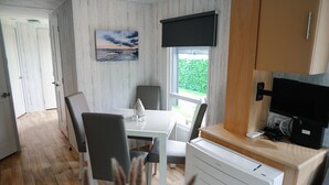 A compact room with a dining table, chairs, a small kitchen area with a TV, and a window with a view of greenery.