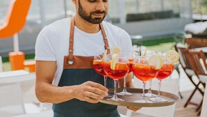 A person in a white shirt and apron carrying a tray with red drinks and lemon slices. - Wabi Hotel - Beauty & Dental Center (Sopron)