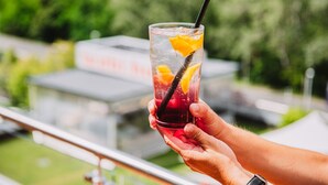 A person holding a glass with a drink and an orange slice, with a blurred outdoor background. - Wabi Hotel - Beauty & Dental Center (Sopron)
