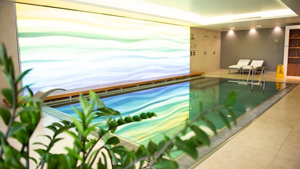 An indoor swimming pool with a wavy wall mural, lounge chairs, and a caution sign. - Wabi Hotel - Beauty & Dental Center (Sopron)