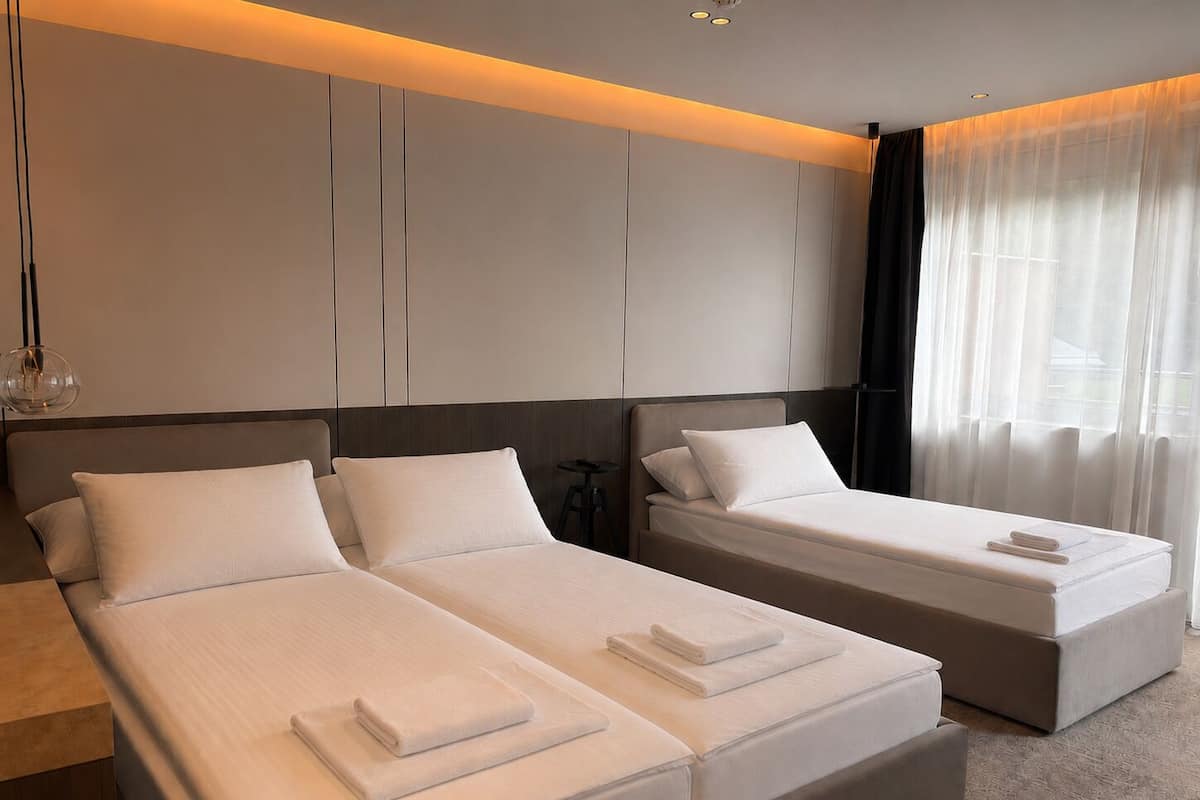A modern hotel room with two beds, a large window with sheer curtains, and a minimalist design.
