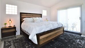 A bedroom with a wooden bed, bedside tables, a floor lamp, and a view of the outside through large windows. - Desert Princess Townhouse Retreat (Cathedral City)