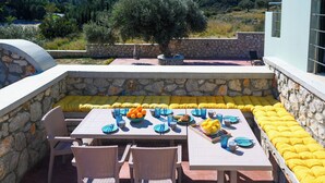 Un espace de restauration en plein air avec des murs en pierre, des coussins jaunes et une table dressée avec des assiettes, des verres et un saladier rempli de fruits.