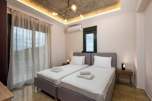 A hotel room with two beds, a wooden nightstand, an air conditioning unit, and a view through a window with curtains.
