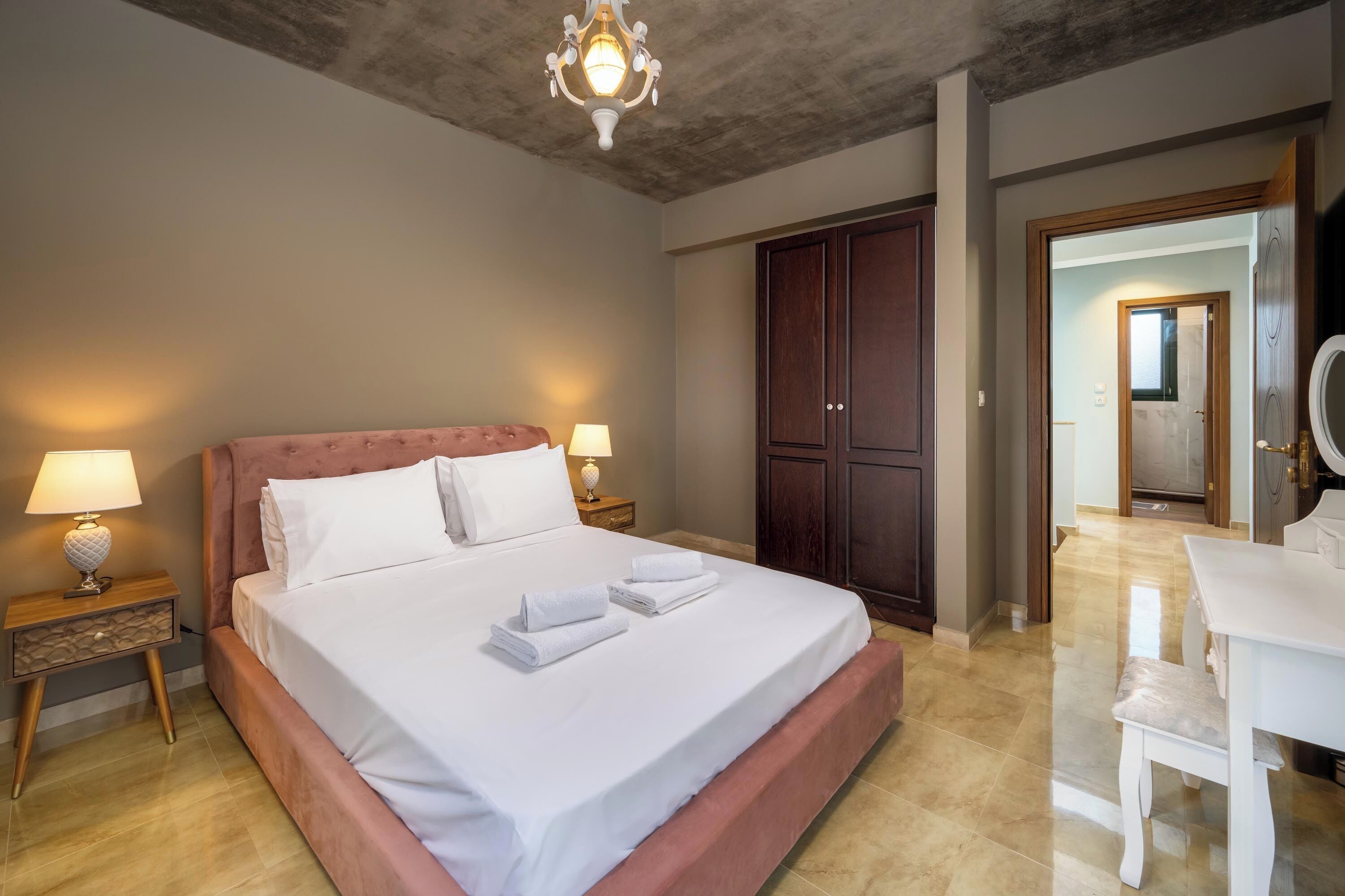Un dormitorio con una cama grande, un armario de madera, mesitas de noche con lámparas, y un baño visible a través de una puerta abierta.