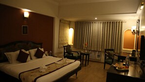 A hotel room with a large bed, a desk, and a chair. There is a window with curtains, a lamp, and a picture on the wall. - HOTEL ANS INTERNATIONAL (Raigarh)