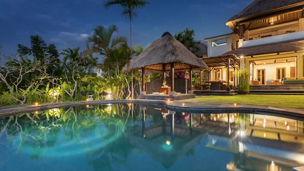 A swimming pool with a thatched-roof gazebo, surrounded by lush greenery and a large house with a balcony. - Villa Djani Balangan by Nagisa Bali (Ungasan)