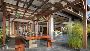 A covered outdoor dining area with a wooden bench and table, surrounded by potted plants and a stone floor. - Villa Djani Balangan by Nagisa Bali (Ungasan)
