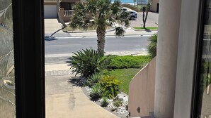 A view from a room with a glass door, overlooking a street with a palm tree, a building with a tiled roof, and a clear blue sky. - OceanView,3500sqft,60sec Beach Access! 4 bedrooms with 4 ensuite baths&Elevator! (South Padre Island)