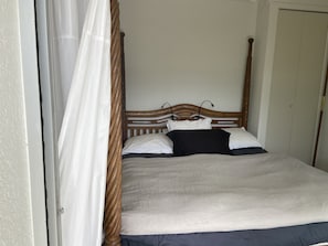 Un lit simple avec une tête de lit en bois, une literie blanche et noire, et un rideau blanc tiré partiellement sur le côté.