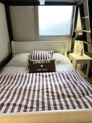 Un lit avec une couvre-lit à rayures, un oreiller orné du texte « LA BAULE Côte Ouest », une lampe et une fenêtre.