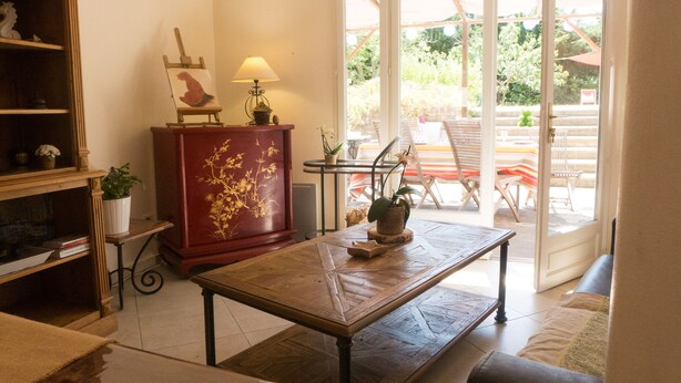 A living room with a wooden coffee table, a red cabinet with floral carvings, a lamp, and a view of an outdoor patio with chairs and a table. - Air conditionned house in a quiet village private pool in large closed garden  (Azille)