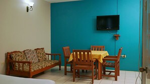A room with a teal wall, a wooden sofa with floral upholstery, a wooden dining table with a yellow tablecloth, and a flat-screen TV mounted on the wall. - King Suite with Kitchenette at Bellissimo (Buccoo)