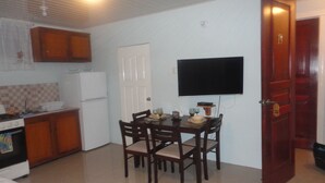 A compact kitchen with a dining area, a flat-screen TV mounted on the wall, and a wooden door with a number plate. - King Suite with Kitchenette at Bellissimo (Buccoo)