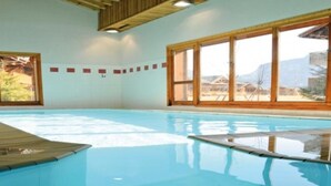 Une piscine intérieure à l’eau d’un bleu cristallin, dotée d’une terrasse en bois et de grandes fenêtres offrant une vue sur l’extérieur.