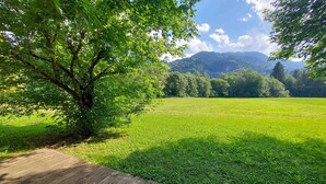 Une terrasse en bois mène à un champ verdoyant, avec un arbre et des montagnes en arrière-plan.