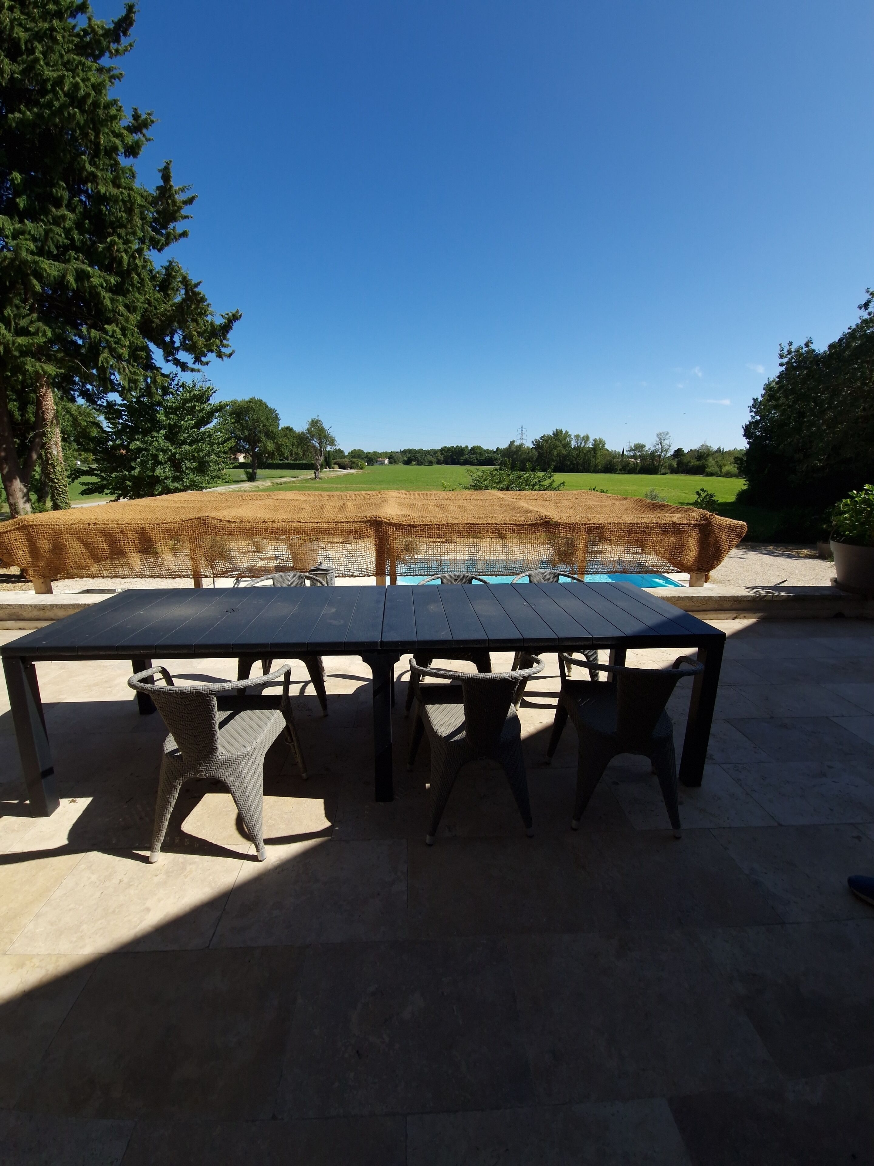 An outdoor dining area with a table and chairs, a pool in the background, and a view of a green field.