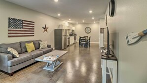 Una sala de estar moderna con un sofá gris, una mesa de centro y un comedor con mesa y sillas. Se ve una cocina con electrodomésticos y un refrigerador al fondo.