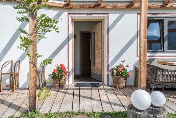 Une terrasse en bois avec une porte donnant sur une pièce, des plantes en pot et un banc.
