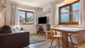 Ein gemütliches Wohnzimmer mit einer Couch, einem Fernseher, einem Esstisch und großen Fenstern mit Vorhängen.
