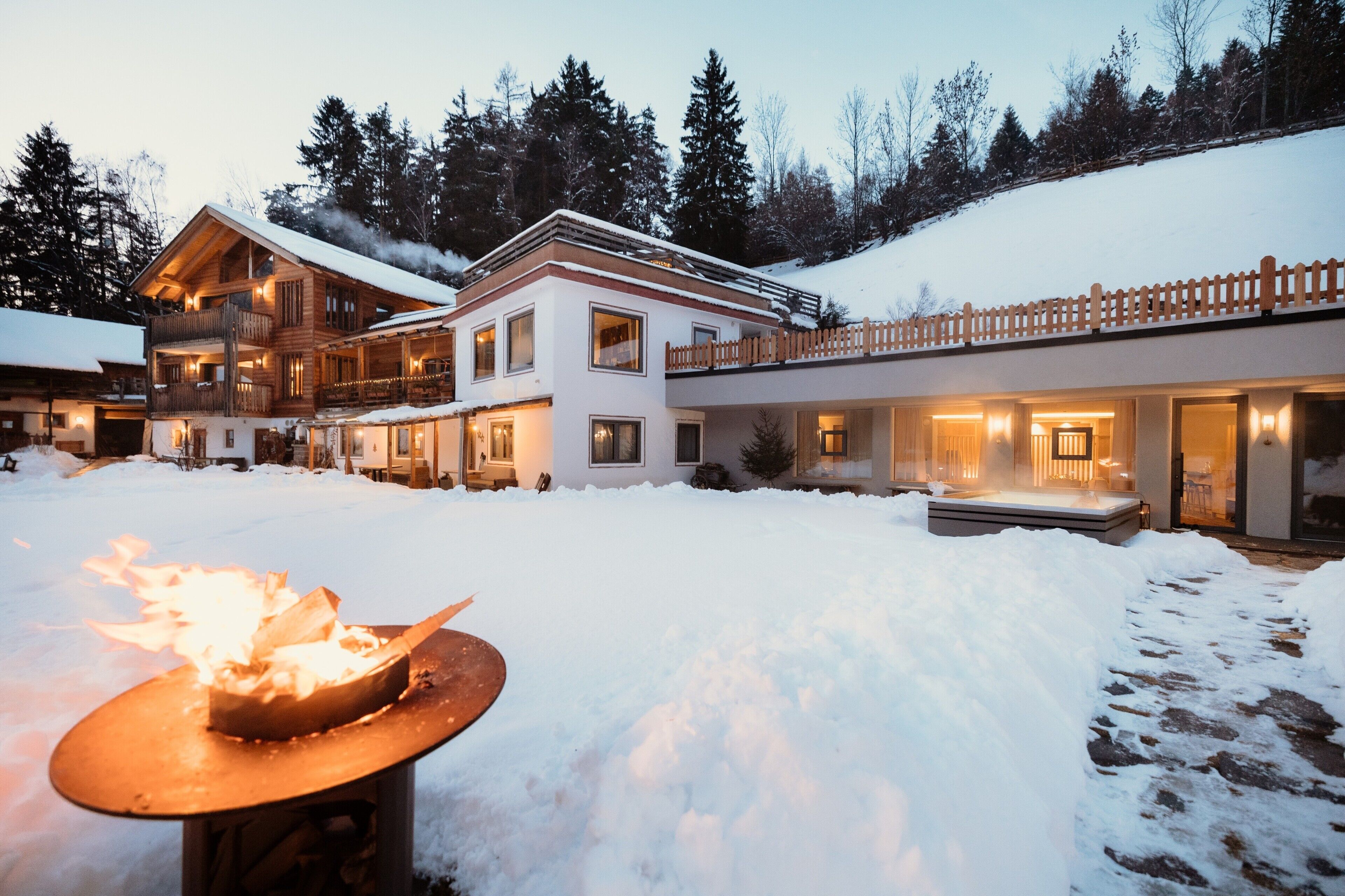 A multi-story chalet with a snow-covered roof, a wooden deck, and a fire pit in the foreground.
