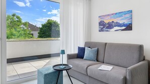 Een moderne woonkamer met een grijze bank, een klein bijzettafeltje en een raam met uitzicht op groen en een gebouw.
