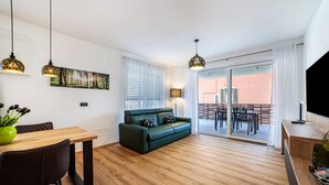 A modern living room with a wooden floor, a green sofa, a dining table with chairs, pendant lights, a painting, and a sliding glass door leading to an outdoor area.