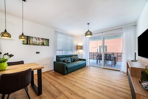 Un salon moderne avec un plancher en bois, un canapé vert, une table à manger avec des chaises, des luminaires suspendus, un tableau et une porte-fenêtre coulissante donnant sur un espace extérieur.