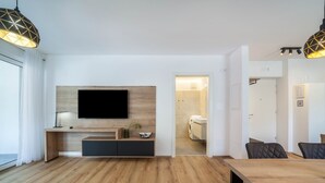 A modern living room with a wooden floor, a flat-screen TV mounted on a wall, a dining area with a wooden table, and a visible laundry room.