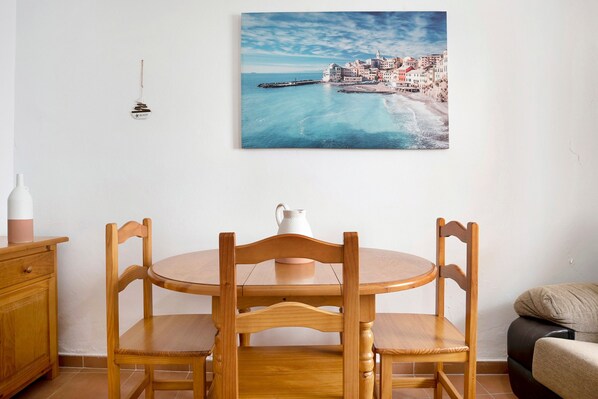 A dining area with a wooden table and chairs, a white pitcher, and a coastal town painting on the wall.