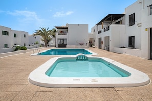 Área de piscina com duas piscinas, edifícios brancos e céu claro.