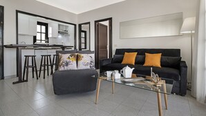 Een moderne woonkamer met een glazen salontafel, een zwarte bank met oranje kussens, een grijze poef en een keuken met witte kasten en een grote spiegel.
