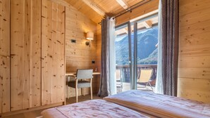 A wooden cabin room with a bed, a desk, and a chair near a window overlooking a mountainous landscape.