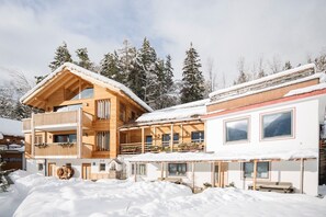 A wooden chalet with a balcony and a covered patio area, surrounded by snow-covered trees.