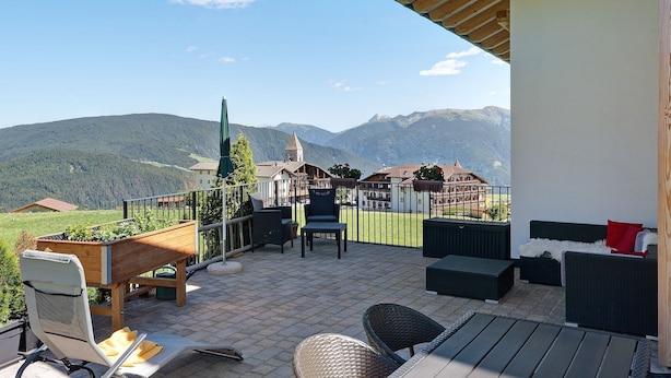 A terrace with outdoor furniture, a wooden planter, and a view of mountains and a church. - Holiday Apartment 'Living Lerchna - Beautiful View' with Mountain View, Balconies and Wi-Fi (Maranza)