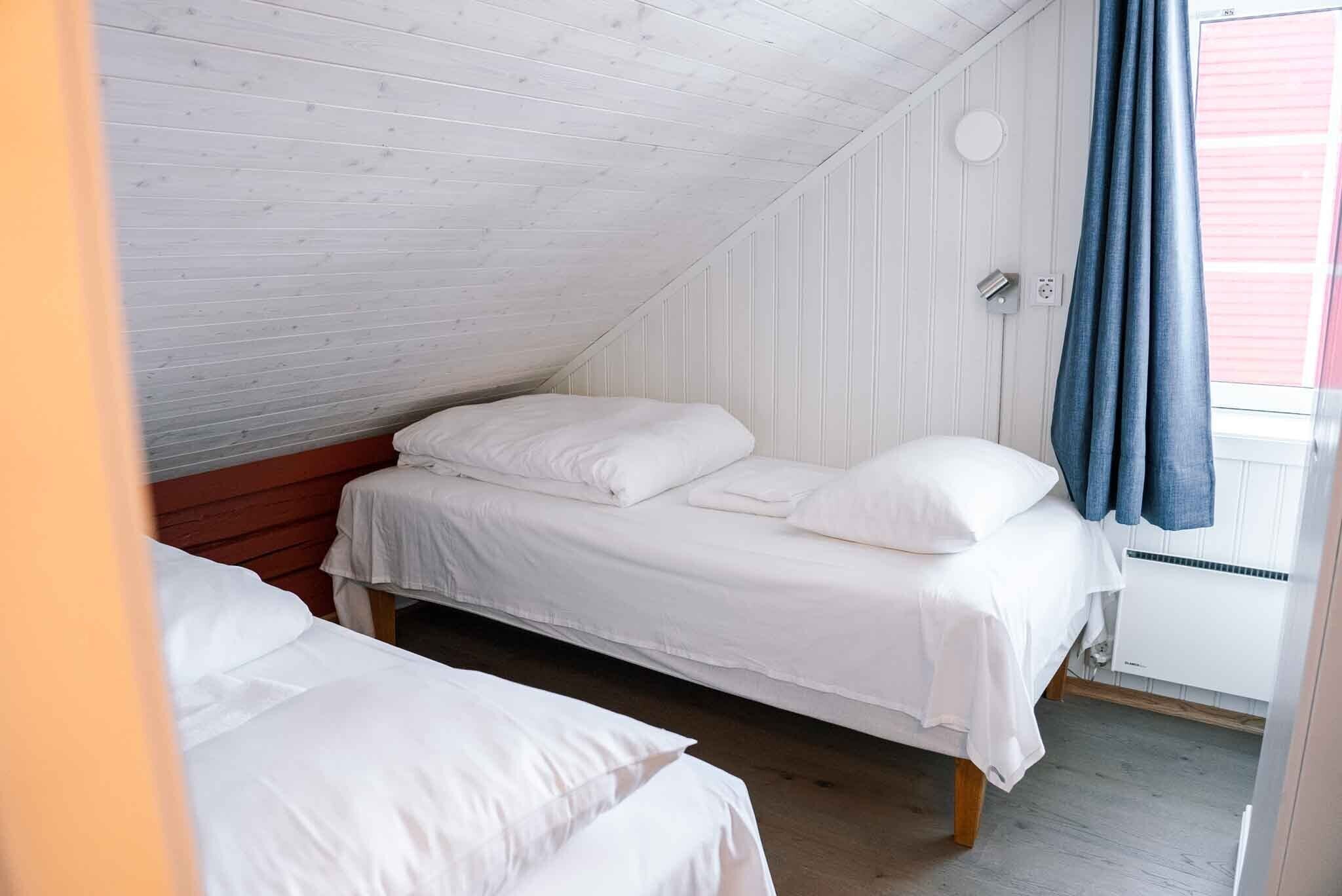 Een knus zolderkamertje met twee eenpersoonsbedden, witte beddengoed, een houten plafond en een raam met een blauw gordijn.