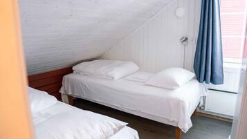 Een knus zolderkamertje met twee eenpersoonsbedden, witte beddengoed, een houten plafond en een raam met een blauw gordijn.