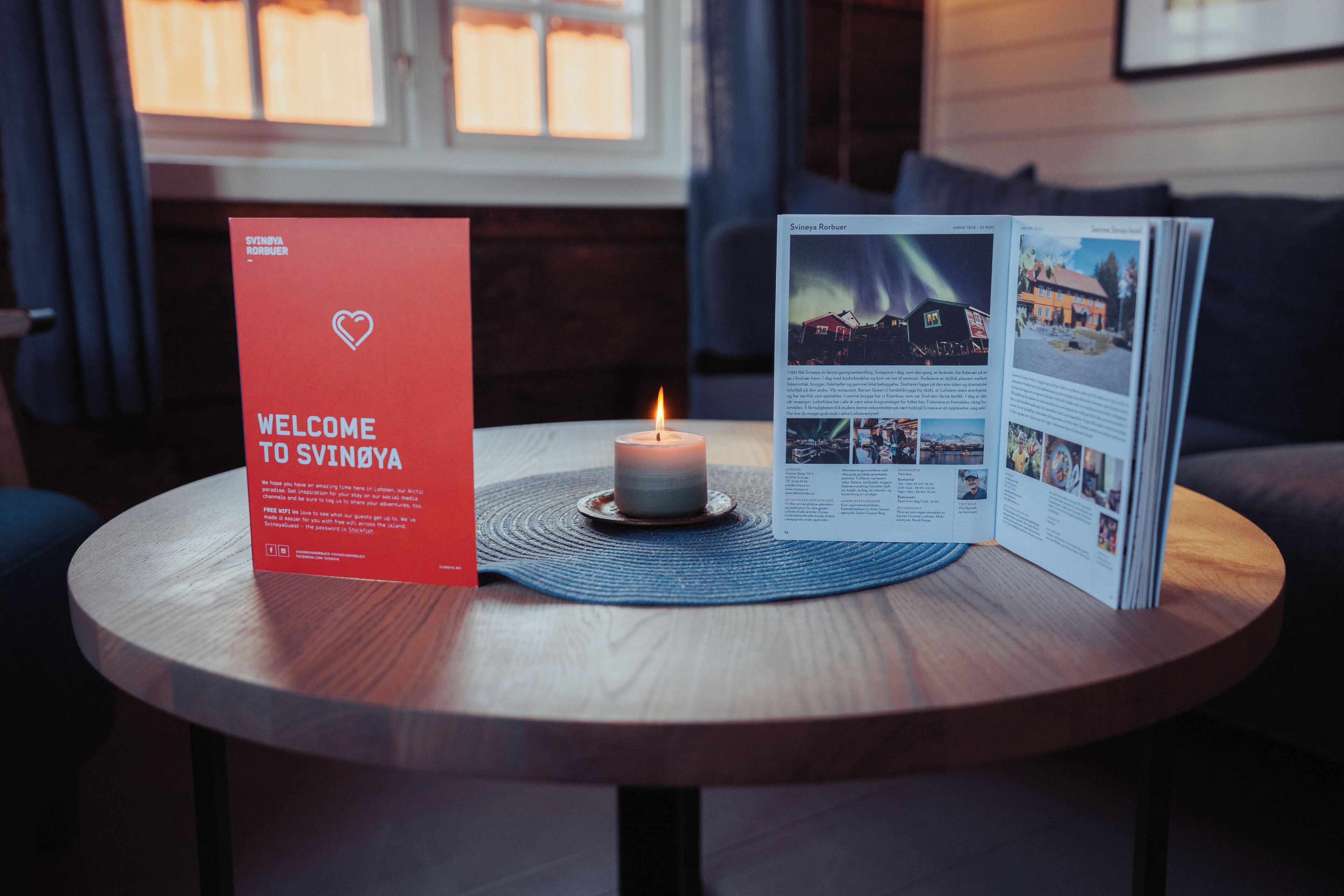 Un panneau d’accueil, une bougie allumée et une brochure posée sur une table ronde en bois.