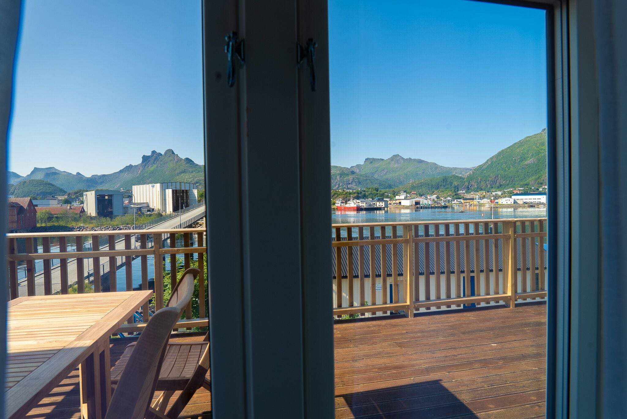 Una terraza de madera con mesa y sillas, con vista a un cuerpo de agua y montañas.
