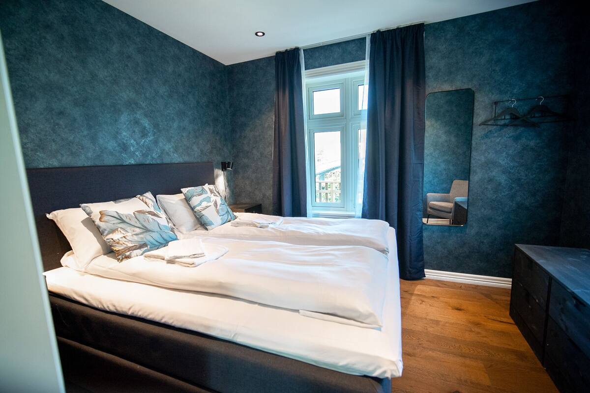 A bedroom with a large bed, a bedside table, a mirror, and a window with curtains.