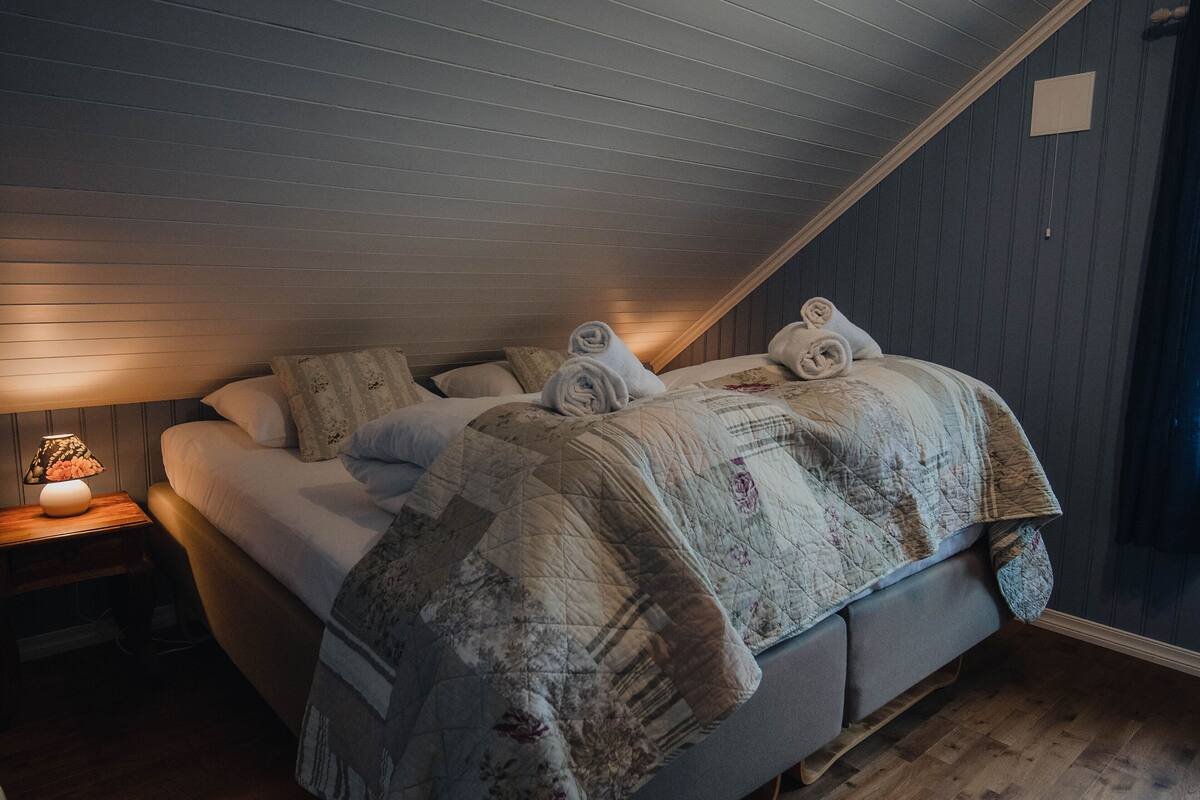 A bedroom with a sloped ceiling, a bed with a quilted bedspread, and a nightstand with a lamp.